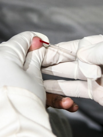 A gloved hand is holding a small lancet near a person's fingertip, with a drop of blood visible on the finger. The focus is on the finger prick procedure.