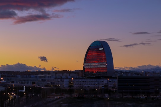 An urban cityscape at dusk with a prominent, uniquely shaped skyscraper featuring reflective glass and modern design elements. The colors of the sunset create a beautiful gradient from deep blue to orange, illuminating the sky. Clouds are scattered across, adding texture to the scene, while city lights begin to glow.