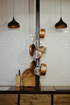 A kitchen scene featuring a hanging rack with a variety of pans and cooking utensils. The background consists of a white tiled wall and two black pendant lights hanging from the ceiling. A wooden countertop displays a knife block and wooden cutting boards.