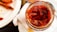 A jar filled with small red chili peppers is placed on a white cloth napkin. Next to it, there is a white plate with a few cooked shrimp. A metallic spoon is partially visible near the jar, adding to the dining context.