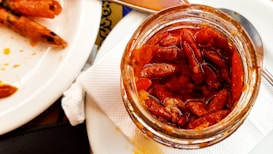 A jar filled with small red chili peppers is placed on a white cloth napkin. Next to it, there is a white plate with a few cooked shrimp. A metallic spoon is partially visible near the jar, adding to the dining context.