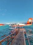 A seaplane docked at a wooden pier floating on a clear blue ocean. The seaplane has a red and white color scheme with the text 'Trans Maldivian Airways' visible. Two people stand near the entrance of the seaplane, while a lifebuoy is seen on the pier. The scene is set under a vast, clear blue sky.