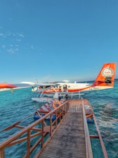 A seaplane docked at a wooden pier floating on a clear blue ocean. The seaplane has a red and white color scheme with the text 'Trans Maldivian Airways' visible. Two people stand near the entrance of the seaplane, while a lifebuoy is seen on the pier. The scene is set under a vast, clear blue sky.