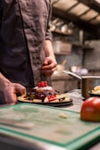 A person in a chef's uniform is preparing a dessert plate filled with pancakes topped with a variety of berries and a dollop of cream. The preparation takes place in a professional kitchen setting, with a blurred background of kitchen equipment and utensils.