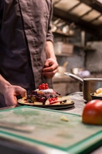 A person in a chef's uniform is preparing a dessert plate filled with pancakes topped with a variety of berries and a dollop of cream. The preparation takes place in a professional kitchen setting, with a blurred background of kitchen equipment and utensils.