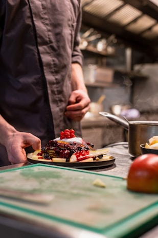 A person in a chef's uniform is preparing a dessert plate filled with pancakes topped with a variety of berries and a dollop of cream. The preparation takes place in a professional kitchen setting, with a blurred background of kitchen equipment and utensils.