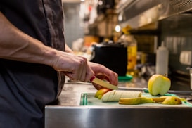 A person is slicing fruit on a cutting board in a kitchen. The fruits include apples and bananas, and are neatly arranged on the board. The kitchen counter is made of metal, reflecting the ambient light. Various kitchen utensils and bottles are visible in the background, suggesting a busy cooking environment.