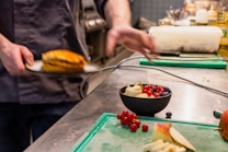 A chef in a kitchen is plating a dish with his hand slightly blurred, indicating motion. A small bowl filled with fresh fruit, including red currants, blueberries, and sliced apples, is placed on the counter alongside multiple chopped ingredients on a green cutting board.