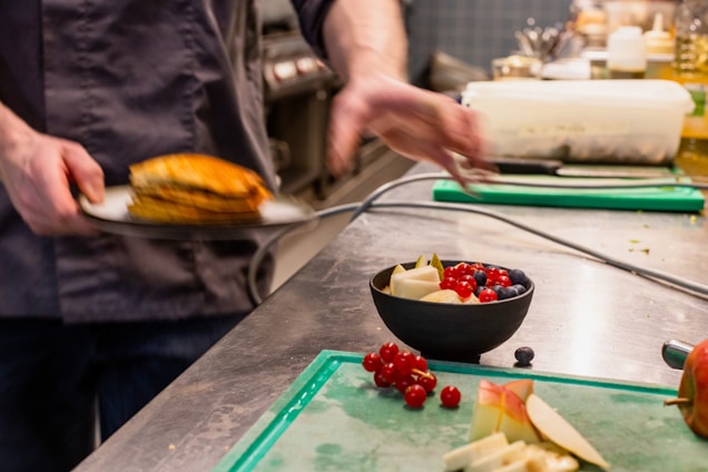 A chef in a kitchen is plating a dish with his hand slightly blurred, indicating motion. A small bowl filled with fresh fruit, including red currants, blueberries, and sliced apples, is placed on the counter alongside multiple chopped ingredients on a green cutting board.