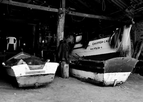 Two boats are stored in a dimly lit indoor setting. The scene has a rustic and weathered look, with visible ropes and fishing nets on the smaller boat. A white plastic chair is set against the background, partially obscured by shadows, and there are wooden beams and structures overhead.