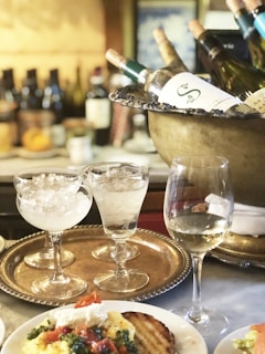 A luxurious dining setting featuring a bucket filled with several bottles of wine on ice, accompanied by a tray with elegant glasses containing icy drinks. In the foreground, there is a plate with a finely arranged meal, including grilled bread topped with a garnish, resembling herbs or vegetables, and a dollop of what appears to be cheese. The background shows a variety of blurred bottles, adding to the ambiance of a refined setting.