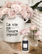 A decorative scene featuring two glass vases and a large ceramic pot filled with light pink roses. The ceramic pot has French text on it, adding a vintage touch. A natural wood surface serves as the base for this arrangement. A woven basket is partially visible in the background.