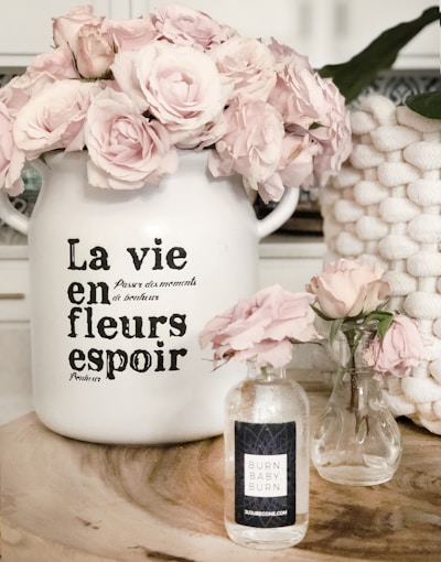 A decorative scene featuring two glass vases and a large ceramic pot filled with light pink roses. The ceramic pot has French text on it, adding a vintage touch. A natural wood surface serves as the base for this arrangement. A woven basket is partially visible in the background.