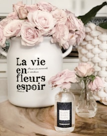 A decorative scene featuring two glass vases and a large ceramic pot filled with light pink roses. The ceramic pot has French text on it, adding a vintage touch. A natural wood surface serves as the base for this arrangement. A woven basket is partially visible in the background.