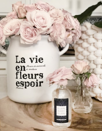 A decorative scene featuring two glass vases and a large ceramic pot filled with light pink roses. The ceramic pot has French text on it, adding a vintage touch. A natural wood surface serves as the base for this arrangement. A woven basket is partially visible in the background.