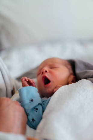 A newborn baby yawns while wrapped in a soft blanket. The infant wears a light blue onesie with small black paw prints. One hand is partially visible, and a caregiver's hand gently holds the baby.