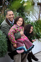 A cheerful parent and child smiling together in a leafy, sunlit playroom filled with natural wood and green plants.