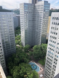 A cluster of tall, modern office buildings surrounds a courtyard area with lush green trees and a small decorative fountain. The buildings are uniformly designed with reflective windows, and the fountain in the center adds an element of tranquility to the urban scene.