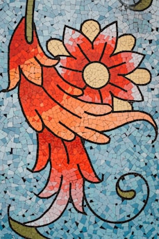 A vibrant mosaic artwork featuring a large, stylized red and orange flower with pointed petals against a backdrop of small light blue tiles. The flower has a yellow circular center and intricate petal details.