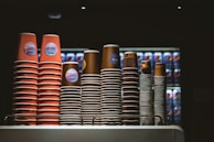 Several stacks of disposable coffee cups are neatly arranged on a countertop. The cups vary in size and color, with some displaying red and others in brown hues. Behind the cups, there is a refrigerator filled with Pepsi cans in a dimly lit environment, suggesting a café or food service setting.