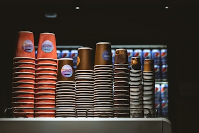 Several stacks of disposable coffee cups are neatly arranged on a countertop. The cups vary in size and color, with some displaying red and others in brown hues. Behind the cups, there is a refrigerator filled with Pepsi cans in a dimly lit environment, suggesting a café or food service setting.