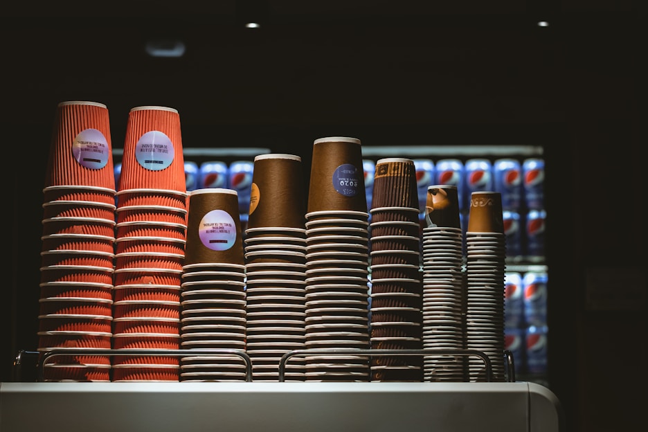Several stacks of disposable coffee cups are neatly arranged on a countertop. The cups vary in size and color, with some displaying red and others in brown hues. Behind the cups, there is a refrigerator filled with Pepsi cans in a dimly lit environment, suggesting a café or food service setting.