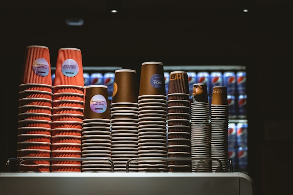 Several stacks of disposable coffee cups are neatly arranged on a countertop. The cups vary in size and color, with some displaying red and others in brown hues. Behind the cups, there is a refrigerator filled with Pepsi cans in a dimly lit environment, suggesting a caf&eacute; or food service setting.