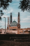 A large building under construction features a prominent teal-colored dome with intricate geometric patterns. The structure is surrounded by scaffolding and various construction materials. Trees partially frame the upper part of the scene, and the sky is clear, suggesting a peaceful environment.