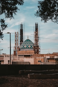 A large building under construction features a prominent teal-colored dome with intricate geometric patterns. The structure is surrounded by scaffolding and various construction materials. Trees partially frame the upper part of the scene, and the sky is clear, suggesting a peaceful environment.