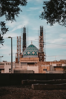 A large building under construction features a prominent teal-colored dome with intricate geometric patterns. The structure is surrounded by scaffolding and various construction materials. Trees partially frame the upper part of the scene, and the sky is clear, suggesting a peaceful environment.