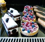 A variety of pastries, including berry-topped tarts with raspberries and blueberries, chocolate and cream cakes with cherries, and glazed éclairs, are displayed on a reflective black surface.
