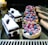 A variety of pastries, including berry-topped tarts with raspberries and blueberries, chocolate and cream cakes with cherries, and glazed éclairs, are displayed on a reflective black surface.