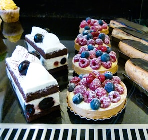 A variety of pastries, including berry-topped tarts with raspberries and blueberries, chocolate and cream cakes with cherries, and glazed éclairs, are displayed on a reflective black surface.
