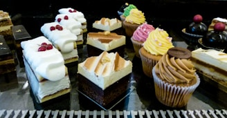 An assortment of pastries and desserts displayed on a reflective surface. The selection includes various cakes, cupcakes with colorful frosting, and layered pastries topped with whipped cream and berries. The desserts are arranged in rows, showcasing a variety of textures and flavors.