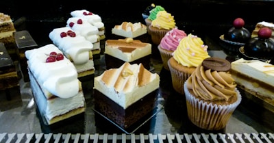 An assortment of pastries and desserts displayed on a reflective surface. The selection includes various cakes, cupcakes with colorful frosting, and layered pastries topped with whipped cream and berries. The desserts are arranged in rows, showcasing a variety of textures and flavors.