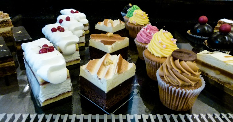 An assortment of pastries and desserts displayed on a reflective surface. The selection includes various cakes, cupcakes with colorful frosting, and layered pastries topped with whipped cream and berries. The desserts are arranged in rows, showcasing a variety of textures and flavors.
