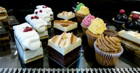 An assortment of pastries and desserts displayed on a reflective surface. The selection includes various cakes, cupcakes with colorful frosting, and layered pastries topped with whipped cream and berries. The desserts are arranged in rows, showcasing a variety of textures and flavors.