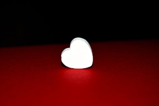 A white heart-shaped object is placed on a rich red surface with a black background. The heart is illuminated, creating a stark contrast with its surroundings.