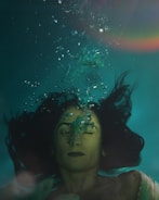 A person with long hair is submerged underwater, with eyes closed and air bubbles escaping from their nose. The surrounding water is a deep teal color, and a subtle rainbow effect is visible in the top right corner.
