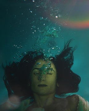 A person with long hair is submerged underwater, with eyes closed and air bubbles escaping from their nose. The surrounding water is a deep teal color, and a subtle rainbow effect is visible in the top right corner.