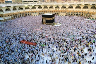A large crowd of people is gathered around the Kaaba in the center of a mosque courtyard. The people are dressed mostly in white, symbolizing a religious gathering. The architecture around the courtyard features arcades and multiple stories filled with people observing the scene.