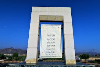 A stone structure with a rectangular frame supports a large marble plaque featuring Arabic script. The surrounding landscape includes a clear blue sky, distant mountains, and vegetation.
