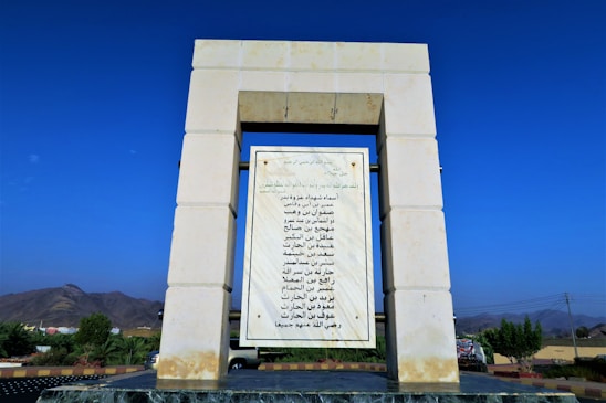 A stone structure with a rectangular frame supports a large marble plaque featuring Arabic script. The surrounding landscape includes a clear blue sky, distant mountains, and vegetation.