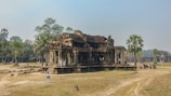 A historic stone temple surrounded by grass and scattered trees. The architecture features intricate carvings and ancient design, with several tourists exploring the site. Nearby, a few palm trees add to the tranquil atmosphere.
