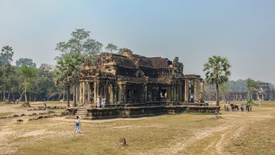 A historic stone temple surrounded by grass and scattered trees. The architecture features intricate carvings and ancient design, with several tourists exploring the site. Nearby, a few palm trees add to the tranquil atmosphere.