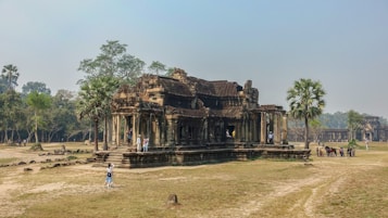 A historic stone temple surrounded by grass and scattered trees. The architecture features intricate carvings and ancient design, with several tourists exploring the site. Nearby, a few palm trees add to the tranquil atmosphere.