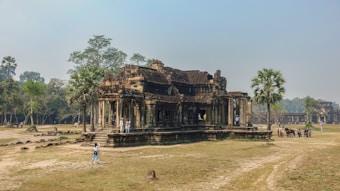 A historic stone temple surrounded by grass and scattered trees. The architecture features intricate carvings and ancient design, with several tourists exploring the site. Nearby, a few palm trees add to the tranquil atmosphere.