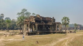 A historic stone temple surrounded by grass and scattered trees. The architecture features intricate carvings and ancient design, with several tourists exploring the site. Nearby, a few palm trees add to the tranquil atmosphere.
