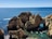 Several boats navigate through turquoise waters around stunning rock formations with arches and cliffs. The horizon shows a vast expanse of calm, deep blue sea, under a clear blue sky.