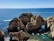 Several boats navigate through turquoise waters around stunning rock formations with arches and cliffs. The horizon shows a vast expanse of calm, deep blue sea, under a clear blue sky.